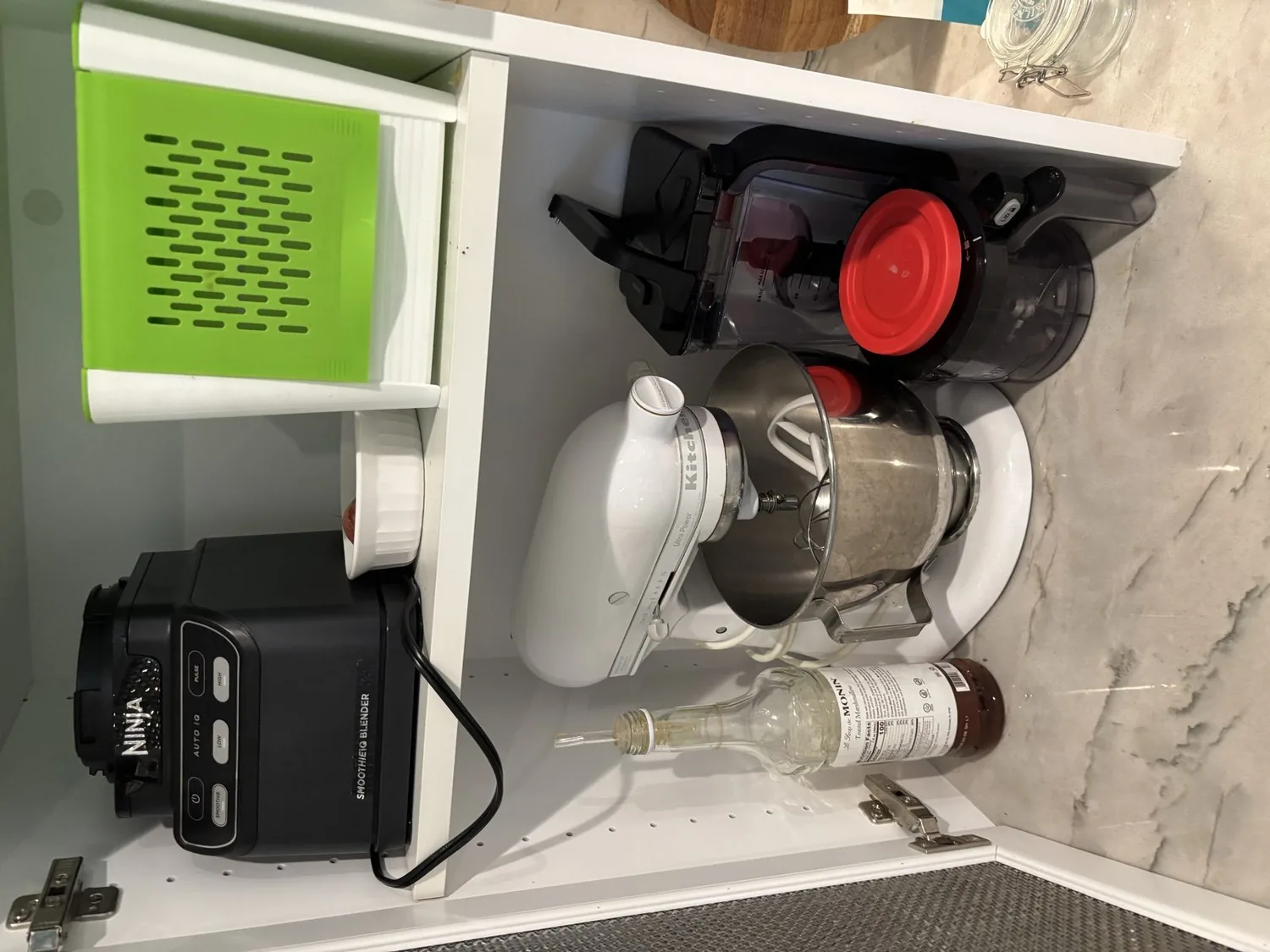 A kitchen cabinet shelf containing a Ninja smoothie blender, a KitchenAid stand mixer, a blender container with a red lid, a green grater, and a bottle of syrup