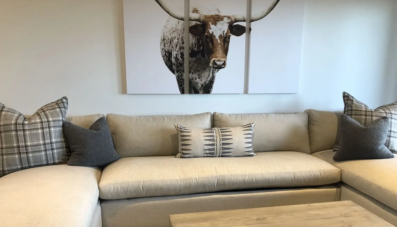 A beige sectional sofa with patterned and solid pillows beneath a three-panel longhorn bull artwork