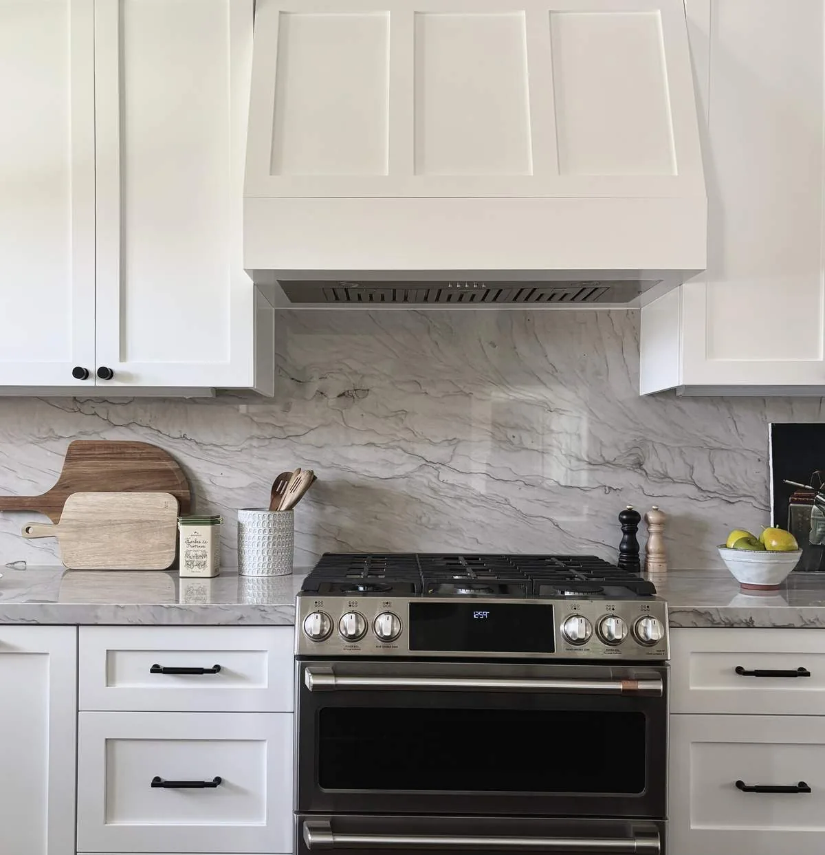 A modern kitchen featuring white cabinetry, a stainless steel stove, marble backsplash and countertop, and wooden cutting boards