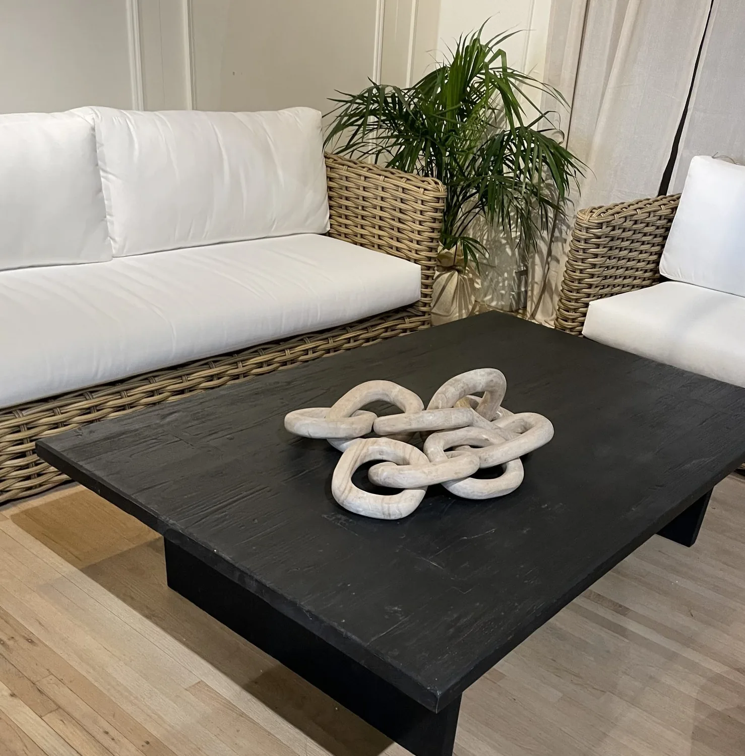 A living room with white cushioned wicker sofas, a black wooden coffee table, and a decorative chain centerpiece