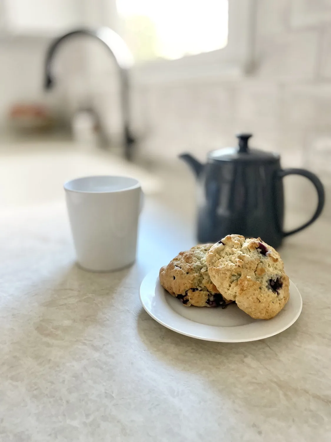 A kitchen countertop with a white cup, a black teapot, and a plate holding two scones