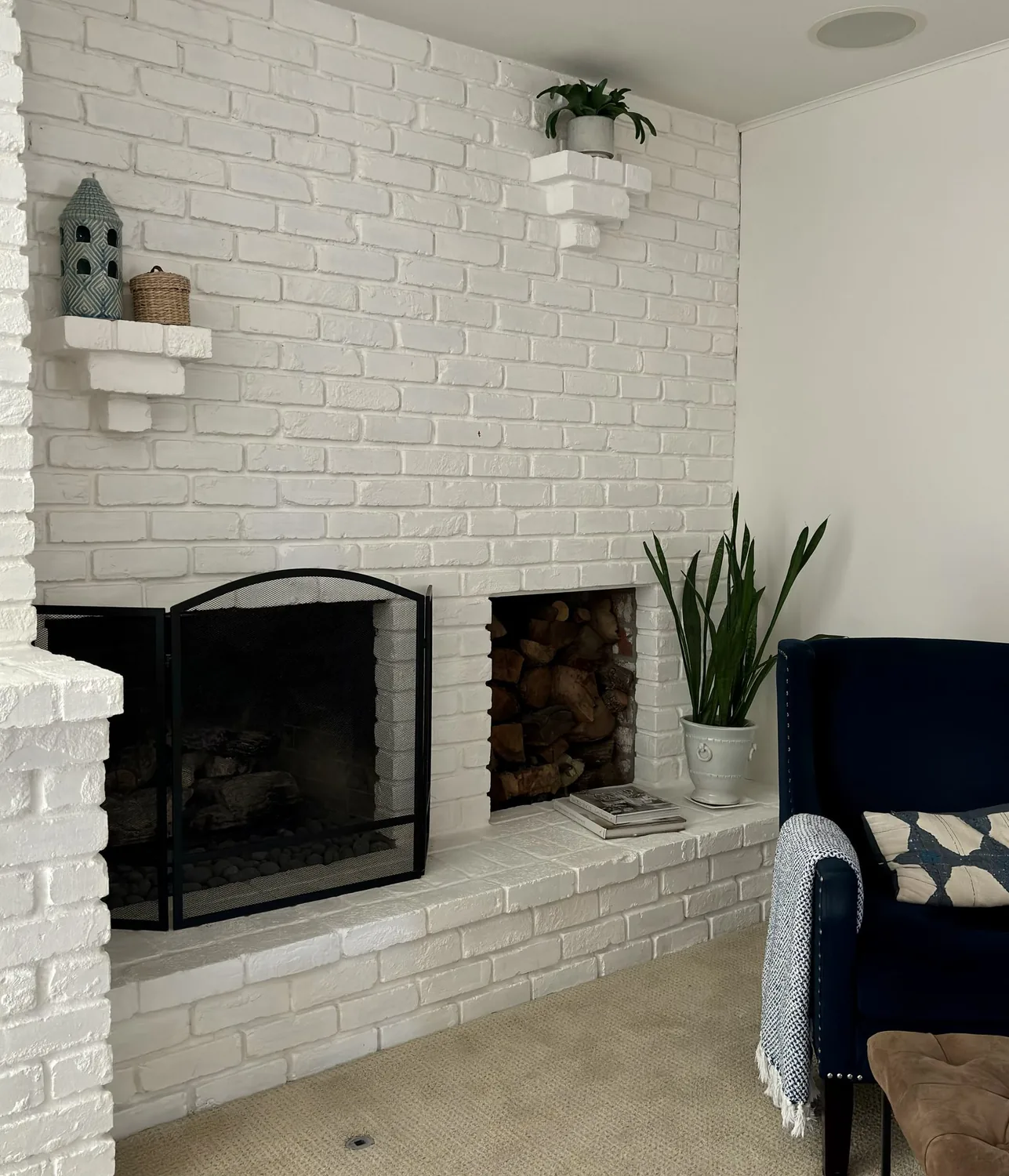 A white painted brick fireplace with a black mesh screen, decorative shelves, a potted plant, and a dark blue armchair