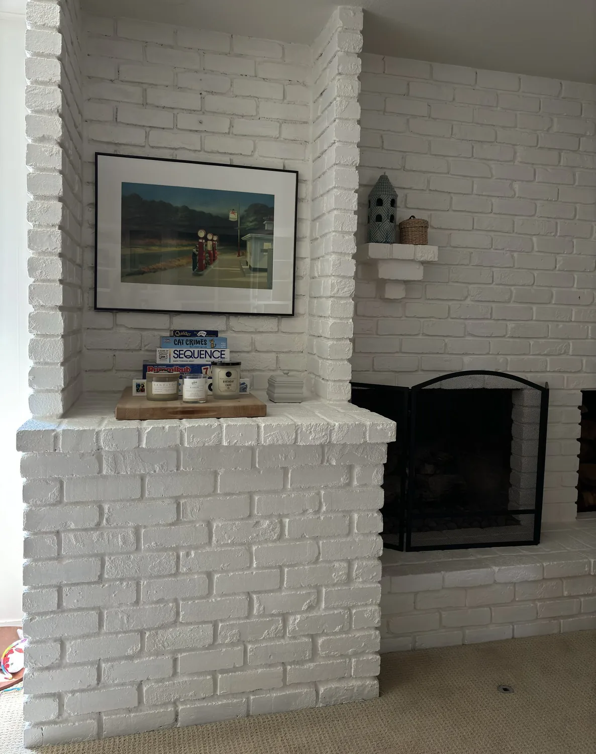 A white painted brick fireplace with a black mesh screen, a framed picture above a ledge holding board games