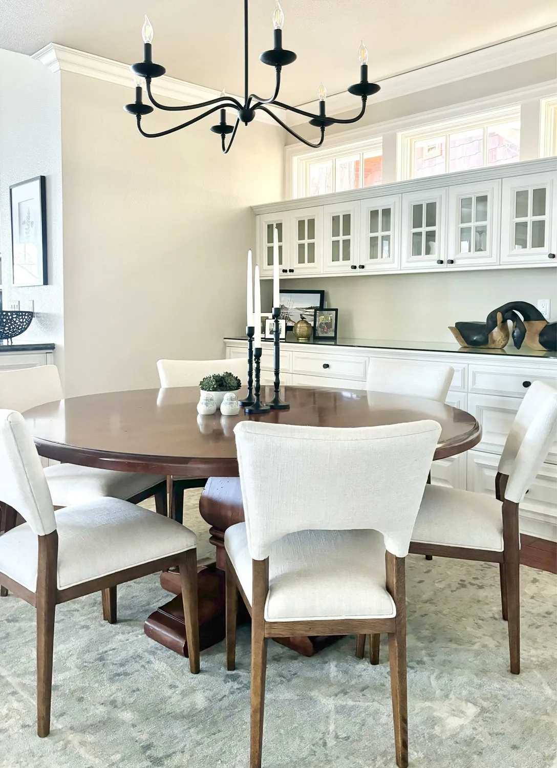 A bright dining room with a round wooden table, six upholstered chairs, a black chandelier, and white cabinetry