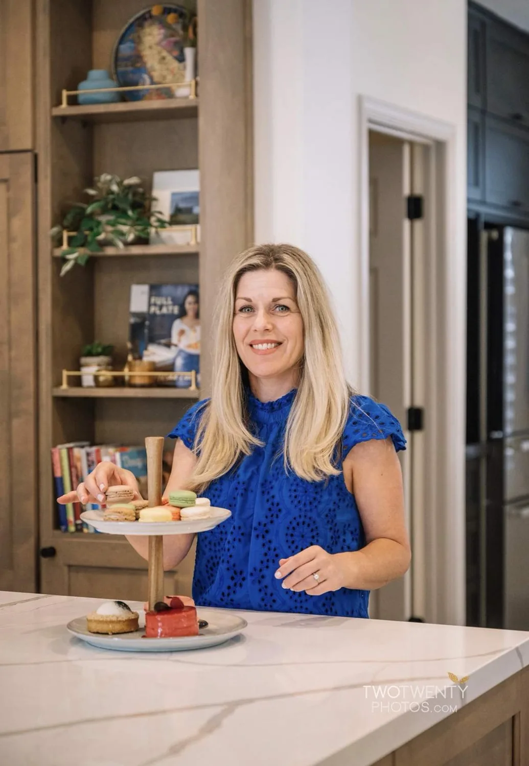 A woman in a blue blouse stands in a modern kitchen holding a tiered tray with macarons and pastries on a marble countertop