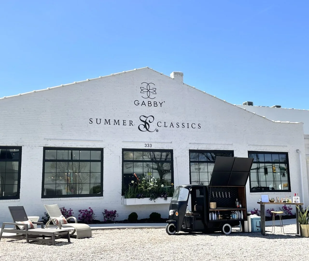 Exterior of a white brick building with black-framed windows, outdoor lounge chairs, and a small black