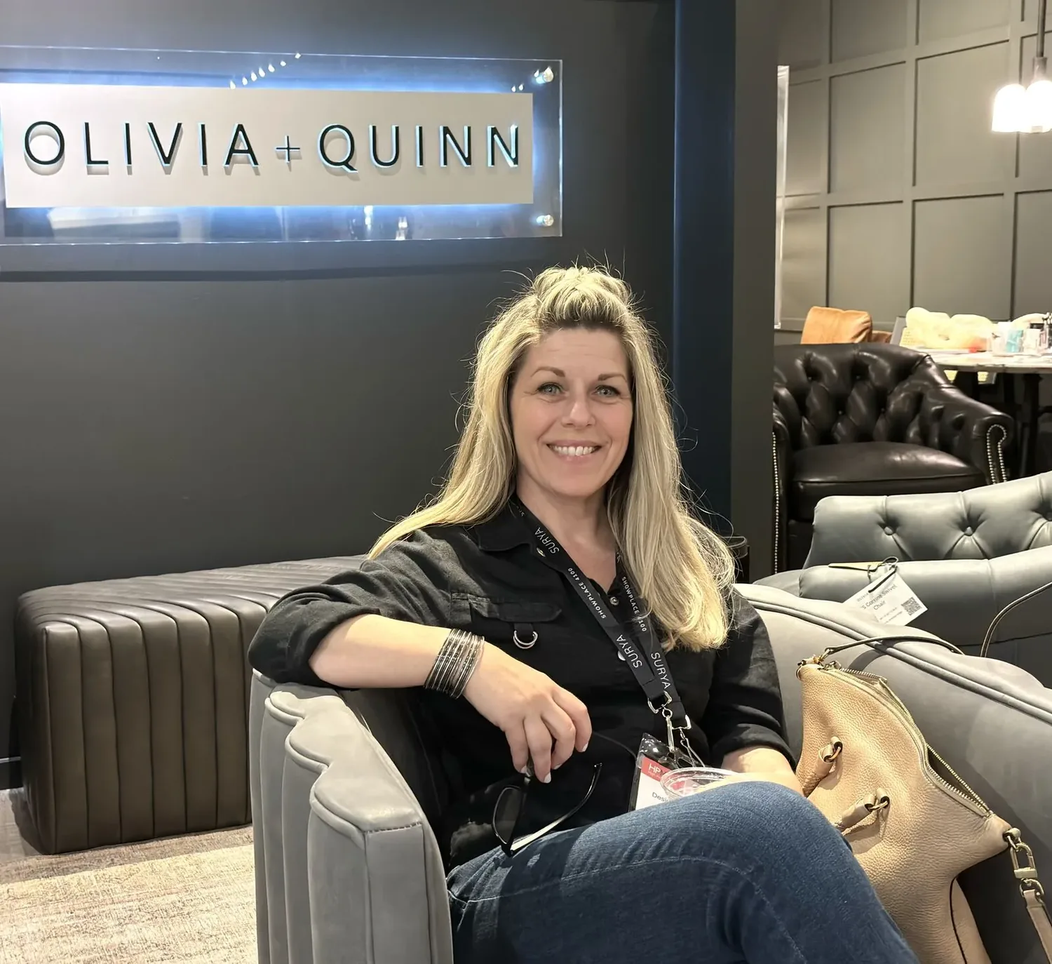 A woman sitting on a gray tufted sofa in a modern showroom with various leather chairs and a sign reading OLIVIA +