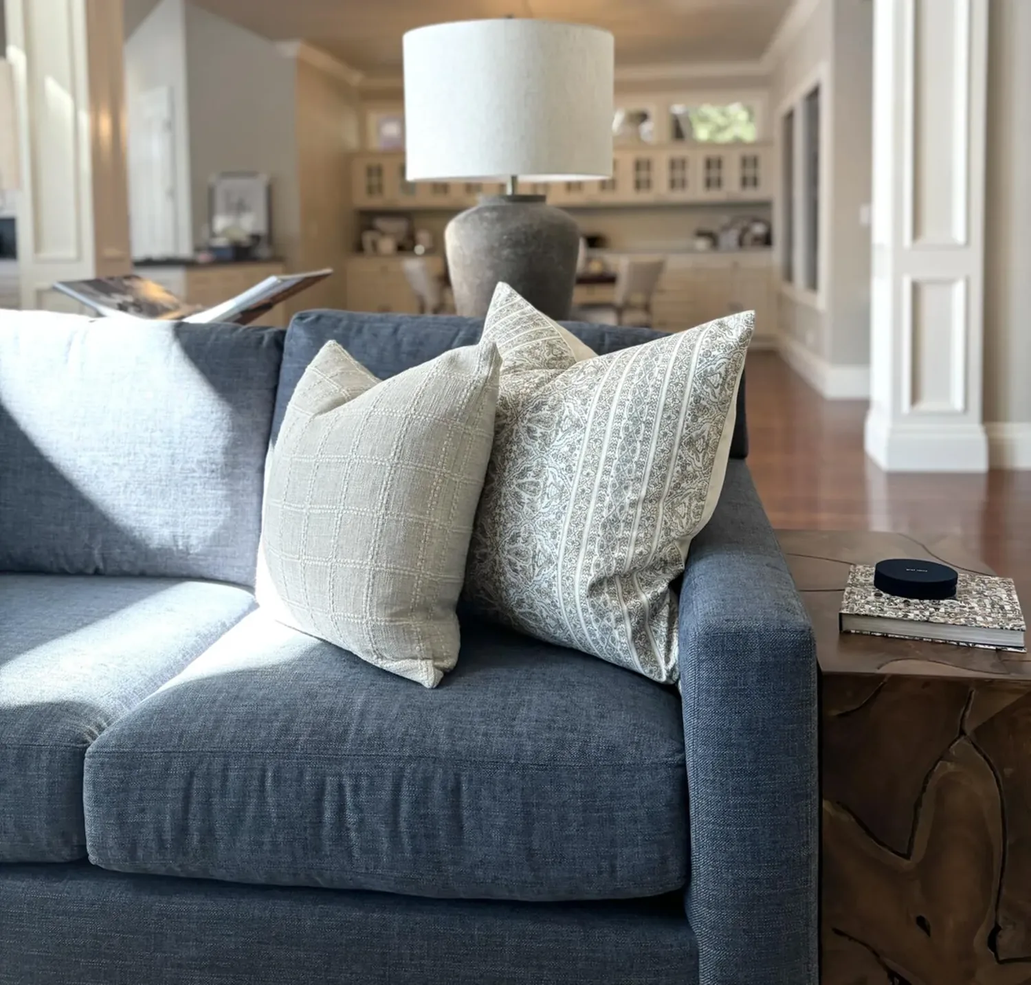 A modern living room with a blue sofa, patterned pillows, a large lamp, and a wooden side table with books and a small
