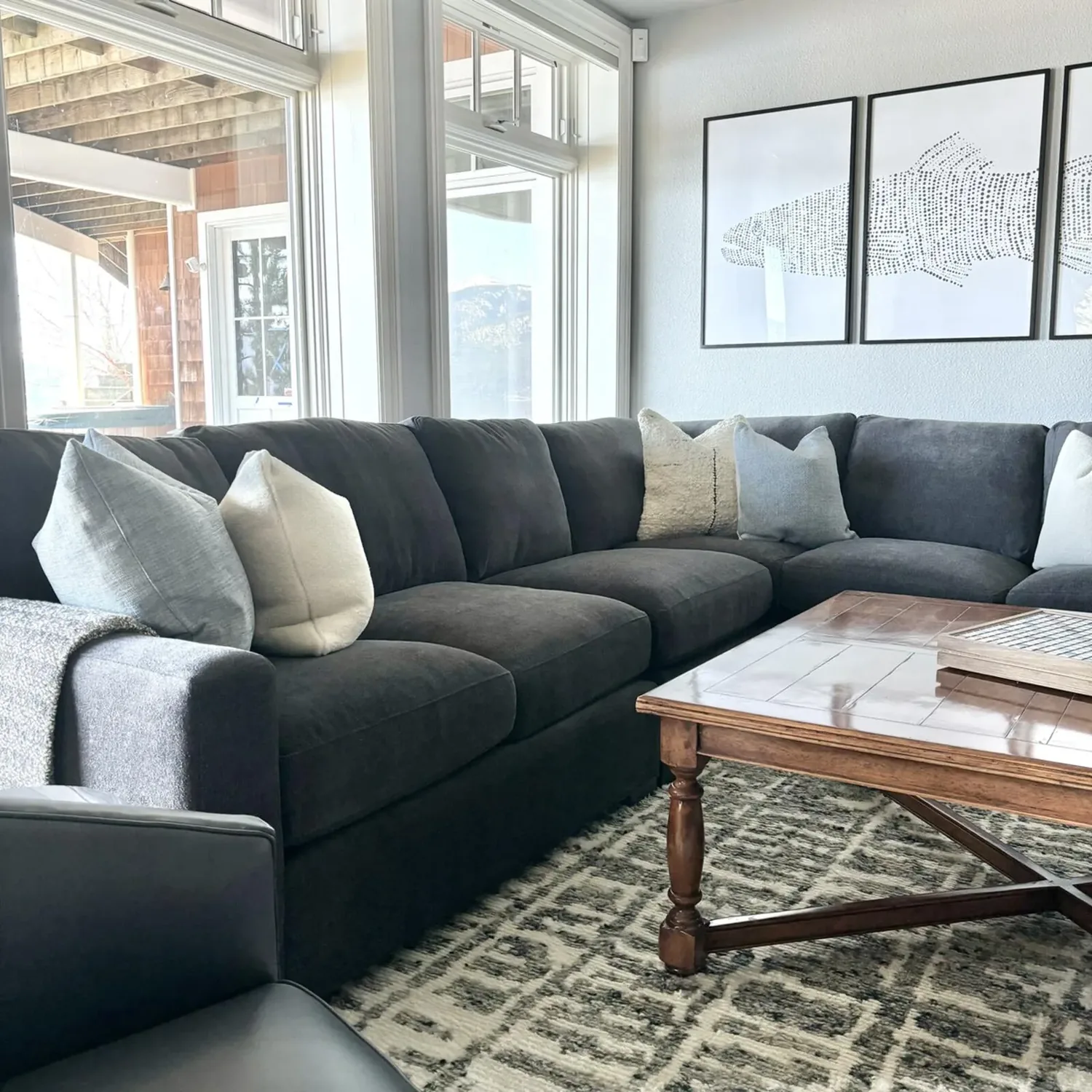 A modern living room with a dark gray sectional sofa, light-colored pillows, a wooden coffee table, and a three-panel fish artwork on the wall.
