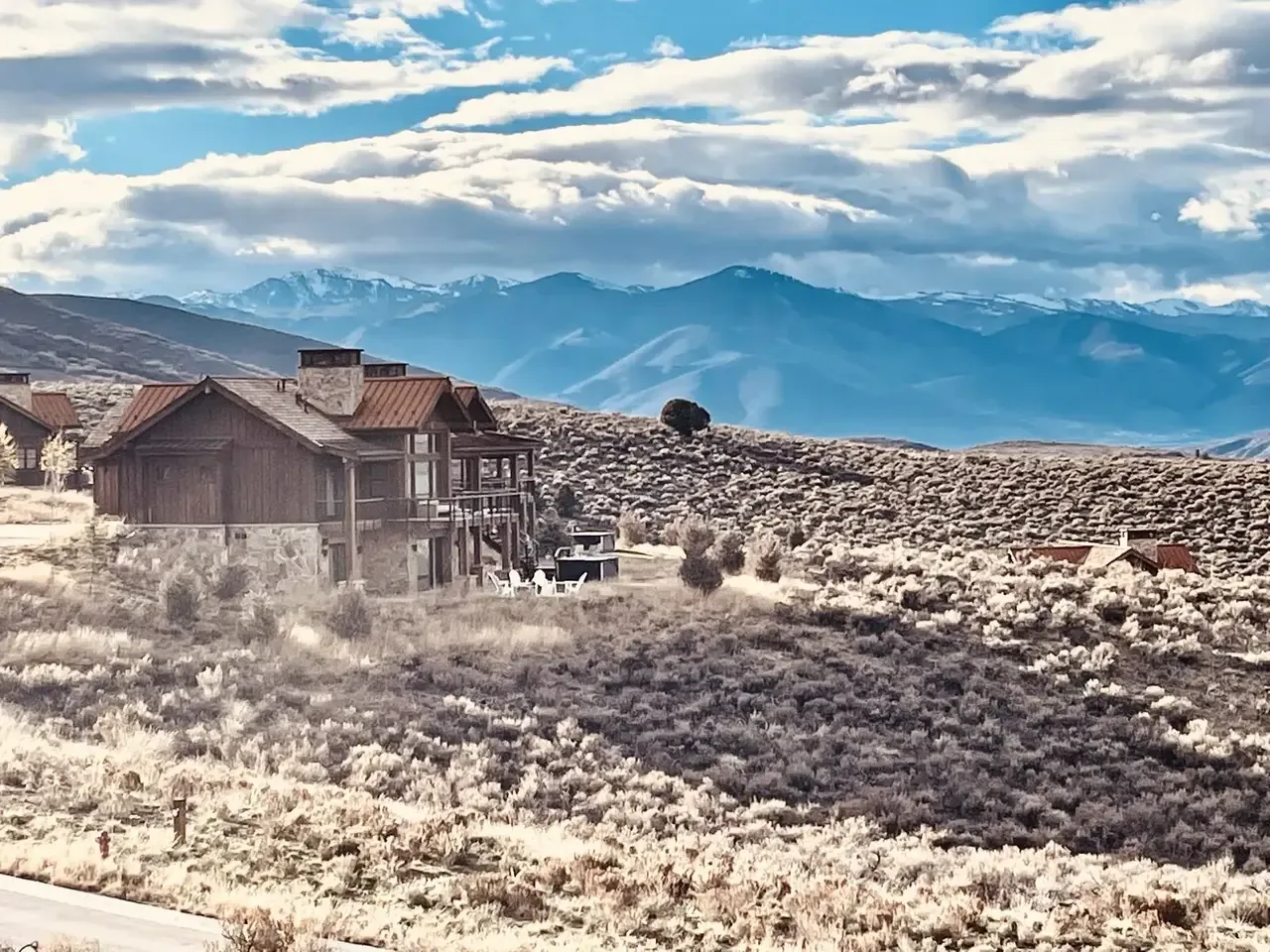 A rustic mountain cabin with stone and wood exterior set in a dry shrubland with mountains in the background