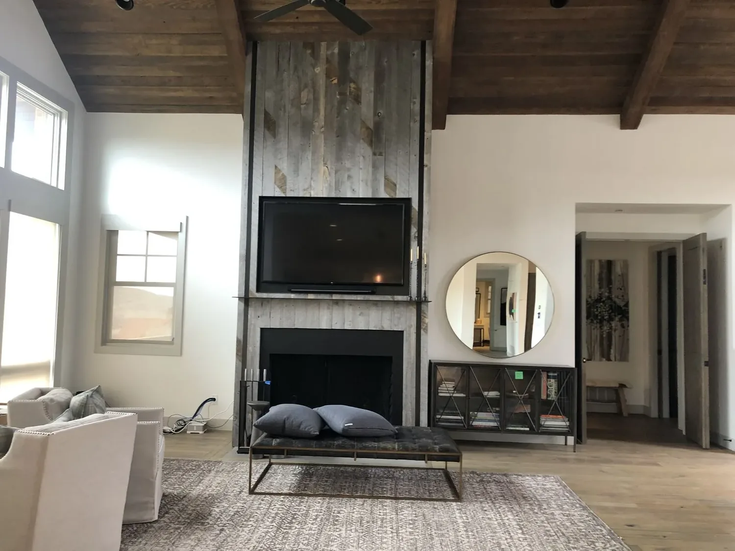 A modern living room with a wooden vaulted ceiling, a TV mounted above a rustic wood-paneled fireplace, a round mirror