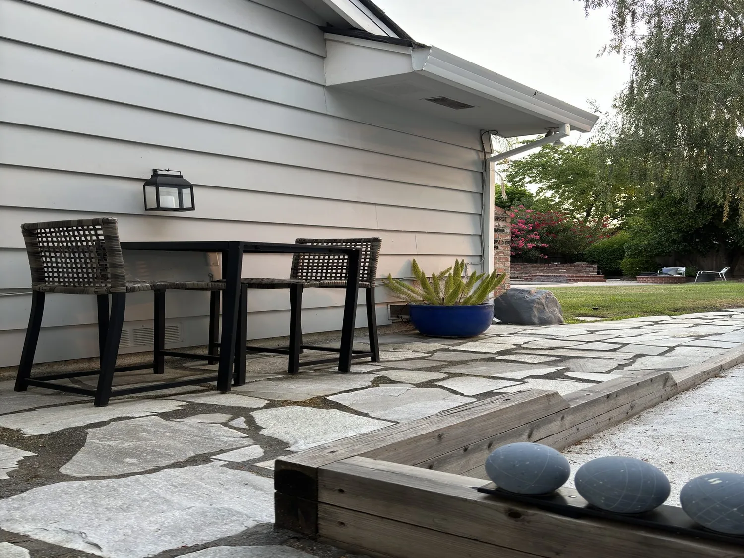 Outdoor patio area with stone flooring, a small table with two woven chairs, a blue planter with greenery, and a bocce