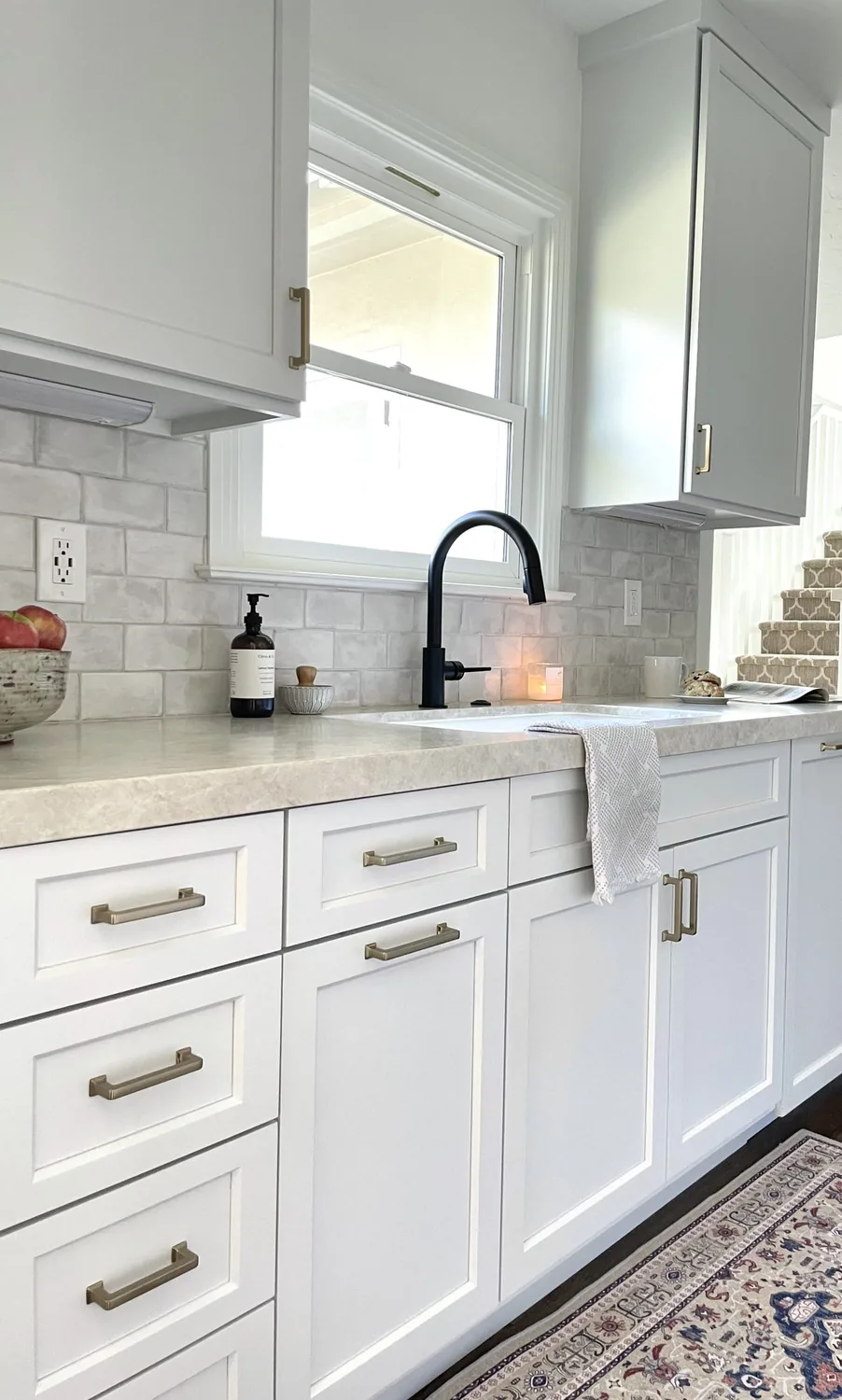 A bright kitchen with white cabinetry, a black faucet, beige countertop, and a patterned rug on the floor
