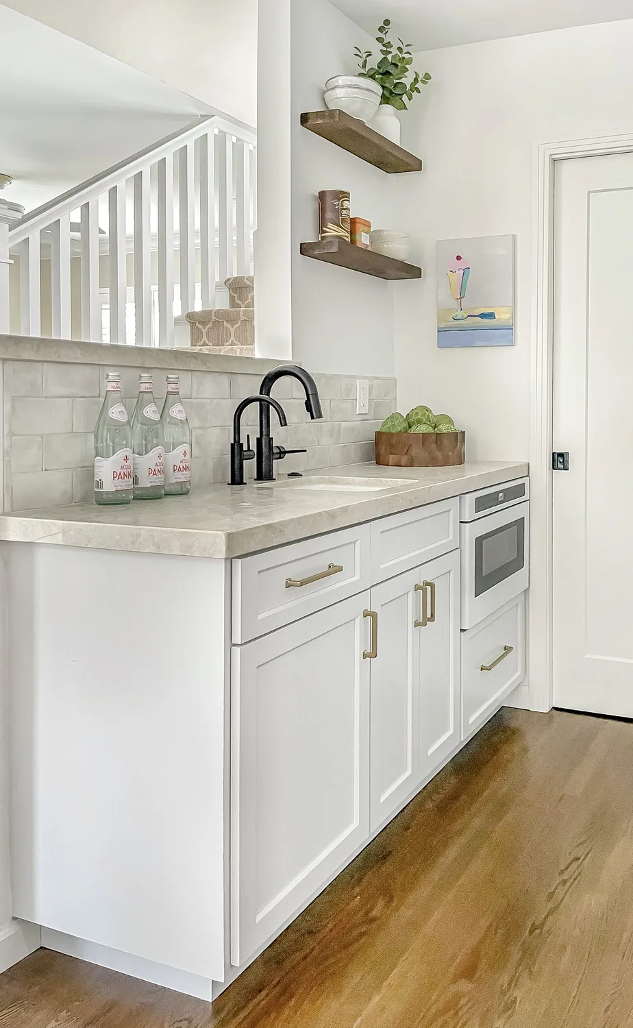 A modern kitchen area with white cabinetry, black faucets, a beige countertop, and decorative shelves on the wall