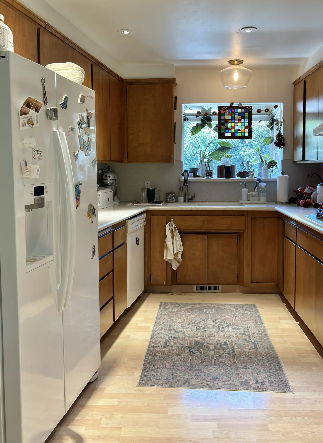 A kitchen with wooden cabinets, a white refrigerator with magnets, a window with plants and a colorful stained glass