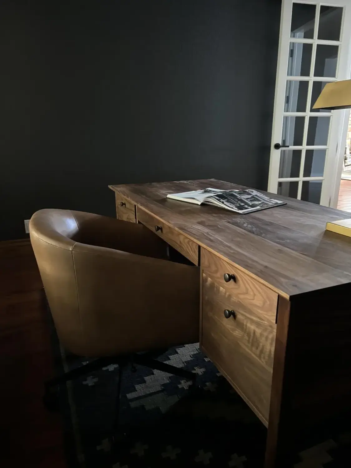 A wooden desk with drawers and a brown leather chair in a dark room with a window and a patterned rug