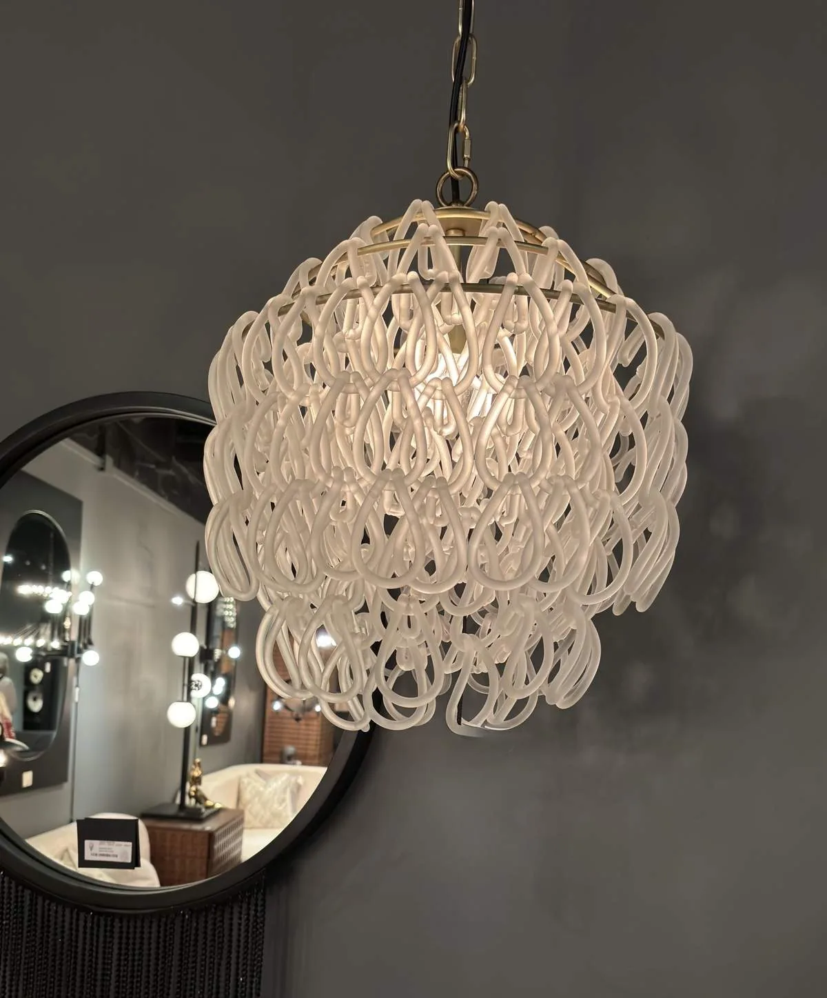 A modern white pendant light fixture with a looped design hanging in front of a round mirror reflecting a room with multiple light bulbs and furniture