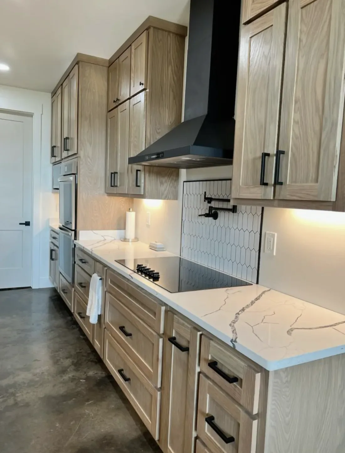 A modern kitchen with light wood cabinets, white marble countertops, a black range hood, and a hexagonal tile backsplash