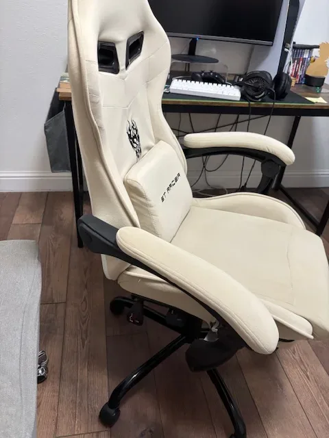 A cream-colored ergonomic gaming/office chair with armrests and lumbar support cushion in front of a desk with computer accessories