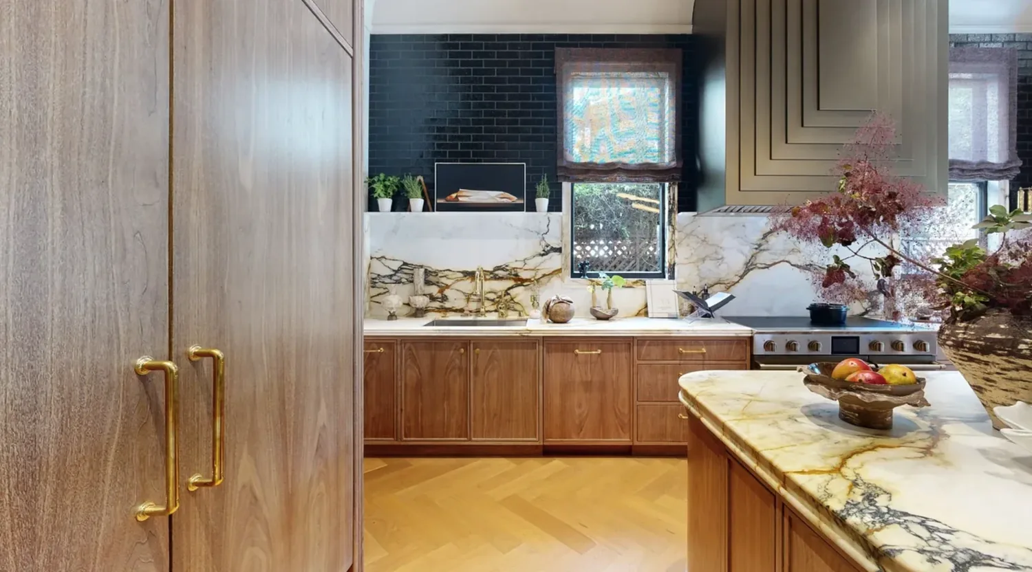 A modern kitchen with wood cabinetry, marble countertops and backsplash, gold hardware, and a black brick accent wall