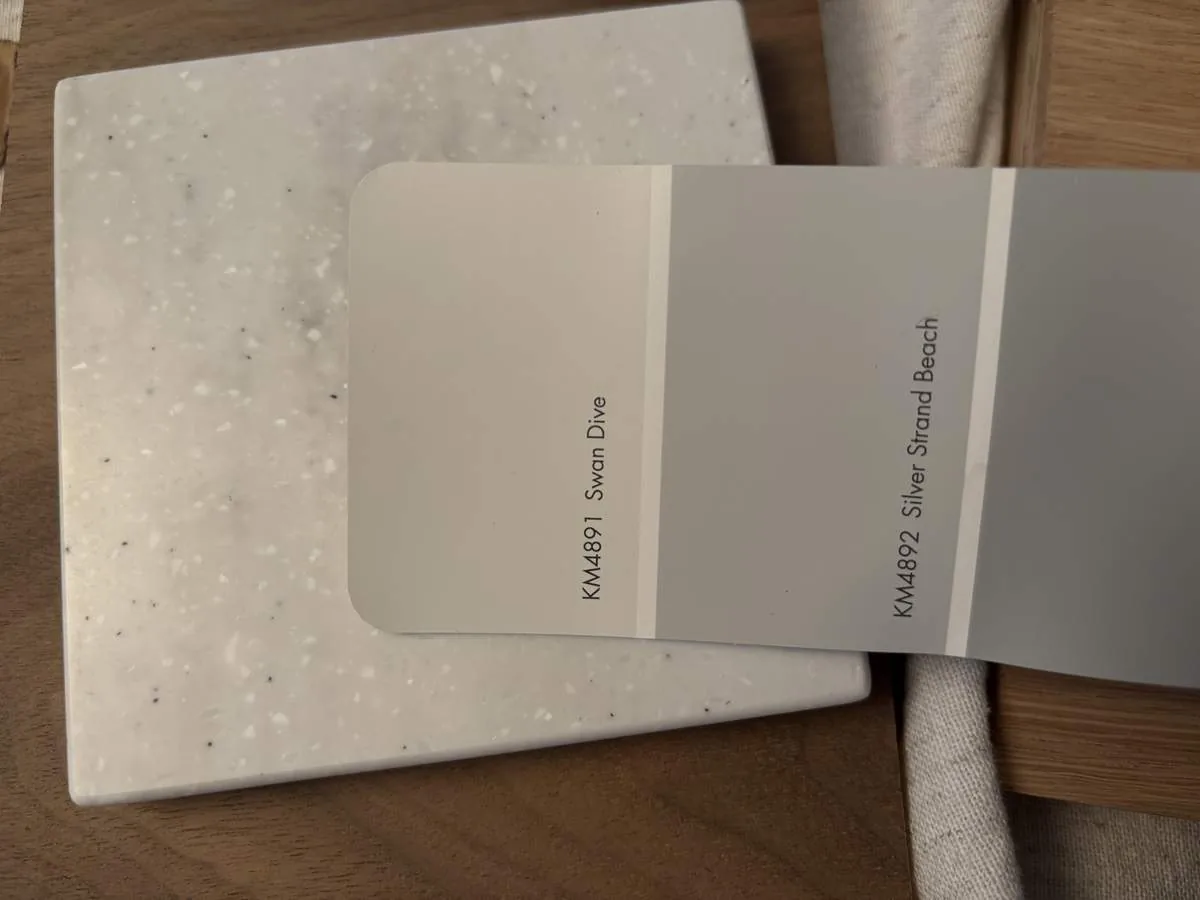 Paint swatches in soft gray tones laid over a white solid-surface sample and oak boards