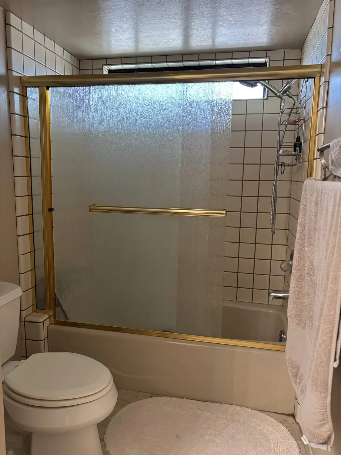 Tiny bathroom. Frosted glass shower, gold trim, white tiles. Beige towels too