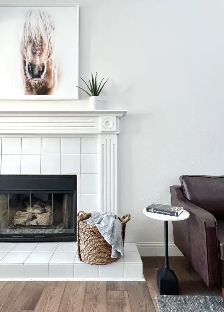 A modern living room corner with a white tiled fireplace, a woven basket with a blanket, a small side table with books