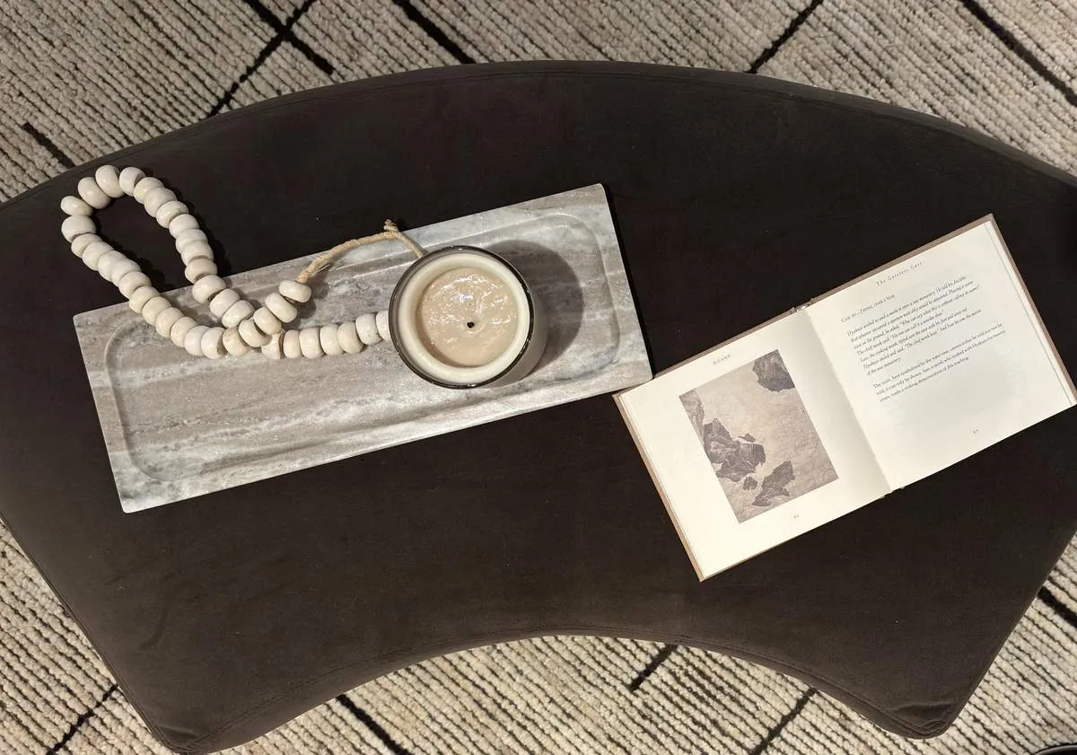 A dark brown curved coffee table on a patterned rug with a marble tray holding a candle and wooden beads, and an open book