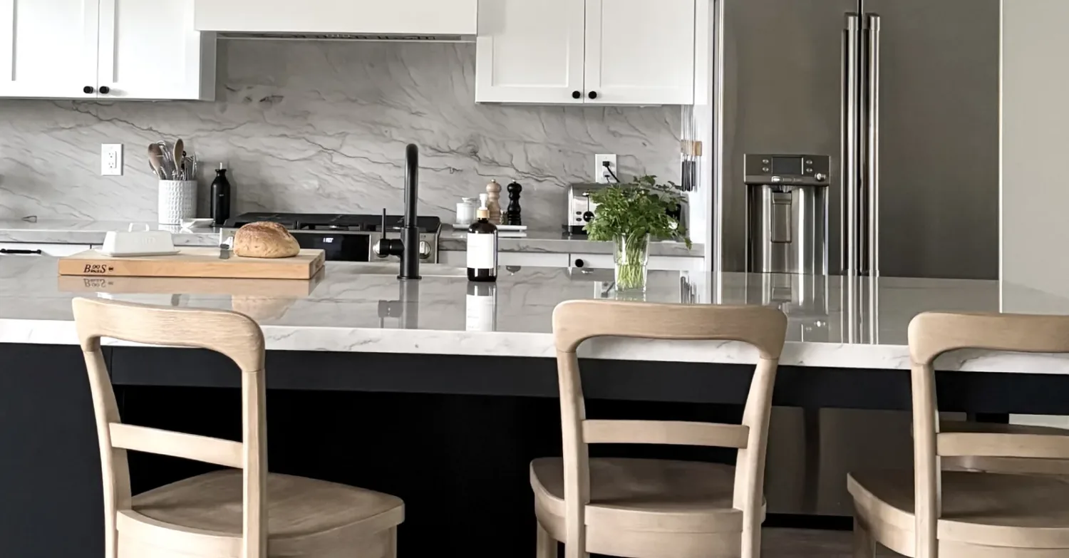 A modern kitchen with a marble countertop island, light wood chairs, white cabinets, and stainless steel appliances