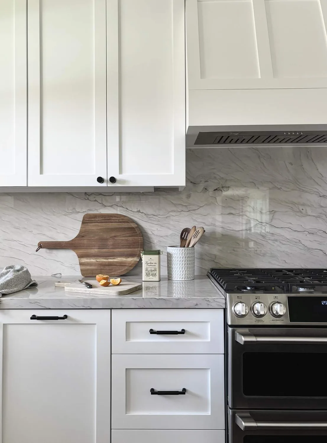 A modern kitchen countertop with white cabinets, a marble backsplash, a wooden cutting board, and a stainless steel stove