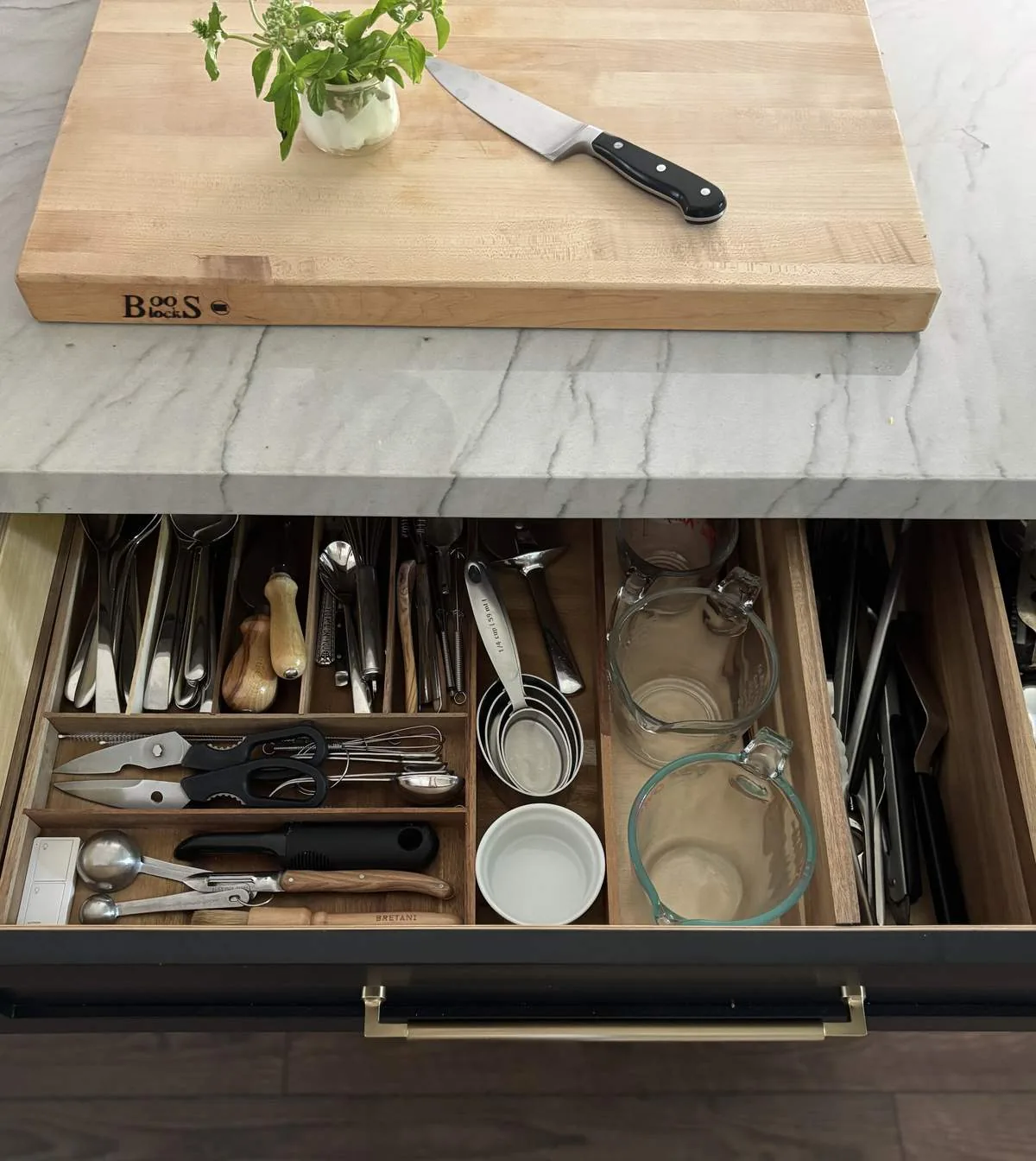 A kitchen drawer organizer with various utensils and measuring cups beneath a marble countertop with a wooden cutting board and knife on top