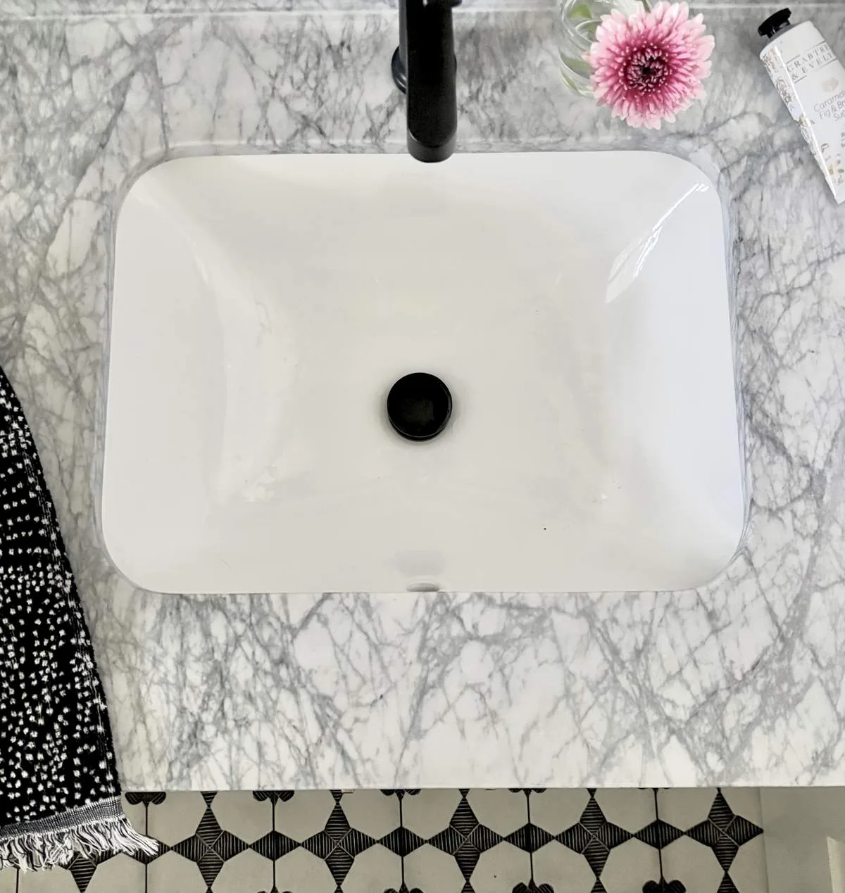 Top-down view of a white rectangular sink set in a gray marble countertop with a black faucet, a pink flower in a glass vase, a black and white towel, and a tube of Crafter & Kelly hand cream