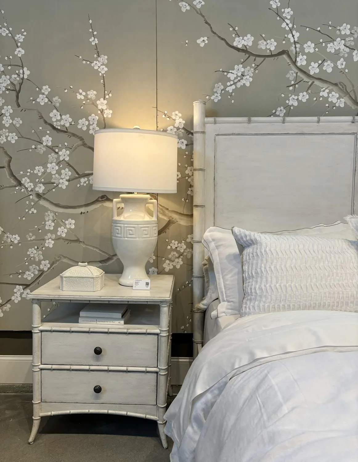 A white bedside table with a ceramic lamp and decorative box next to a white bed with textured pillows, set against a wall with a floral branch mural