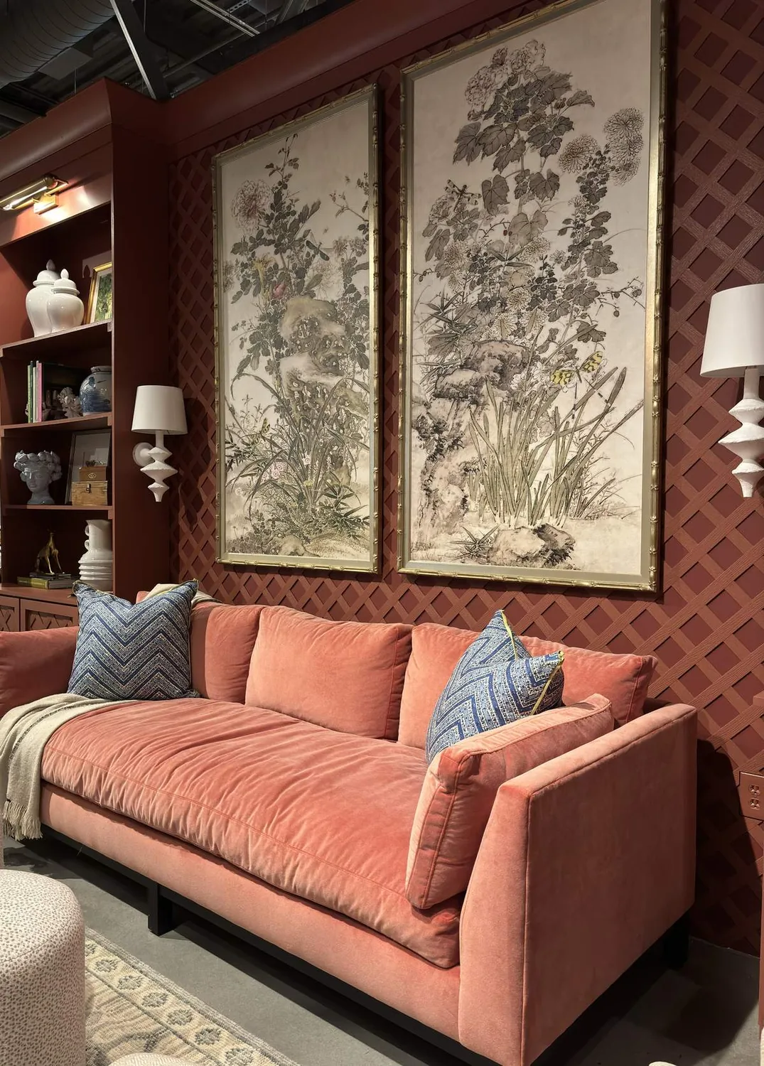 A living room with a coral pink velvet sofa, blue patterned pillows, floral wall art, and a built-in bookshelf against a red lattice wall