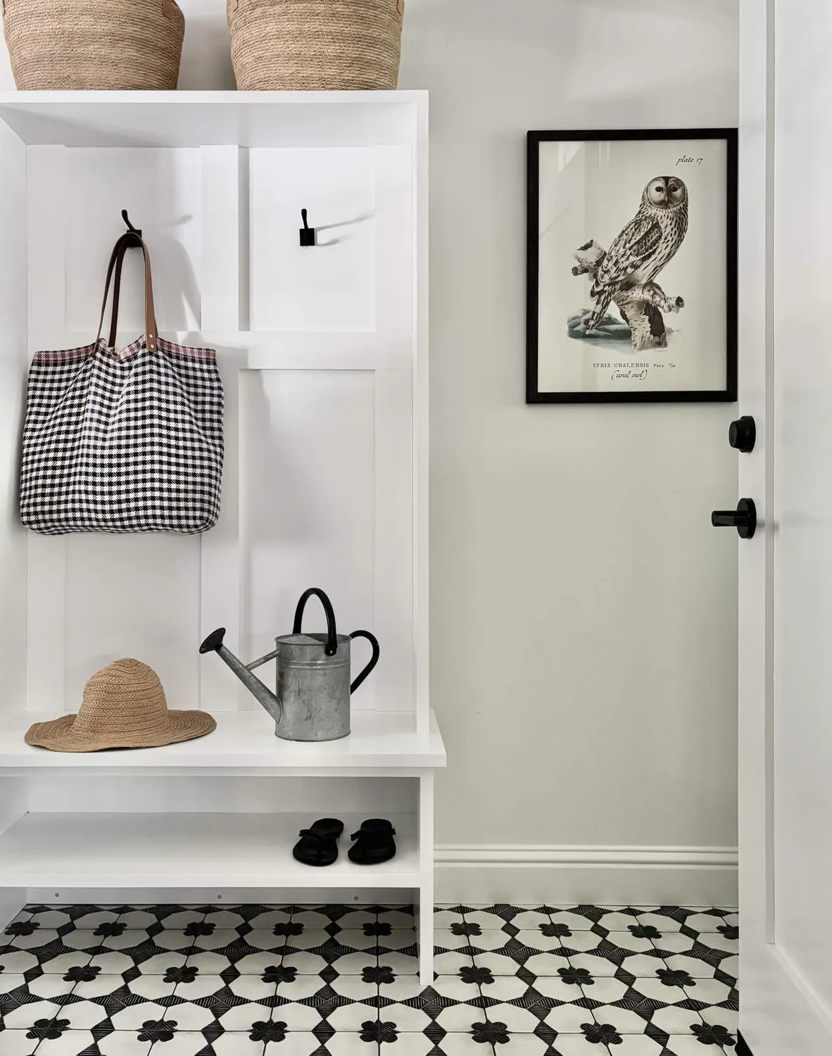 A minimalist entryway with a white built-in bench and hooks, featuring a houndstooth bag, straw hat, watering can