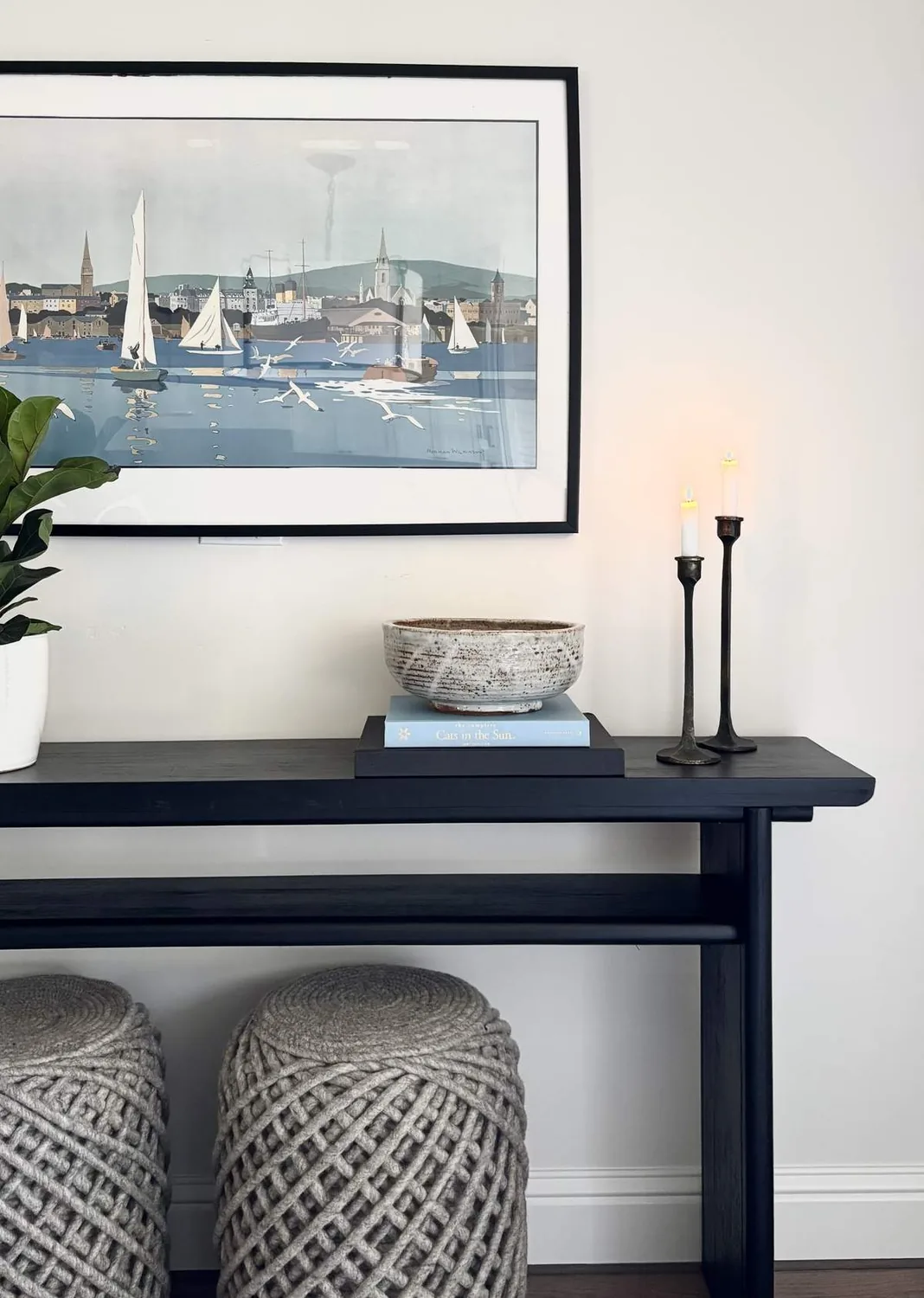 A minimalist styled entryway or living space with a black console table, decorative bowl, two candles, a potted plant, and a framed nautical-themed artwork above