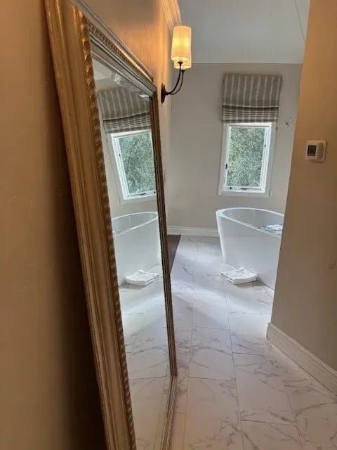 A bathroom with a freestanding bathtub, marble floor tiles, a large ornate mirror, and a wall sconce light fixture