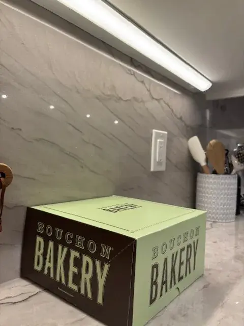 A kitchen countertop with a Bouchon Bakery box, marble backsplash, under-cabinet lighting, and kitchen utensils in a holder