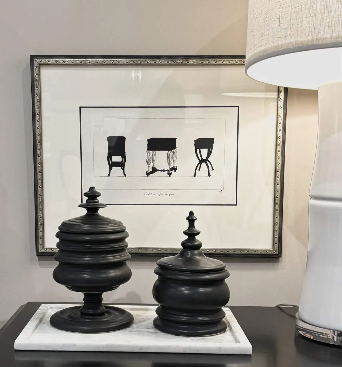 A decorative vignette featuring two black urns on a white tray, a framed black and white print of furniture silhouettes, and a white table lamp