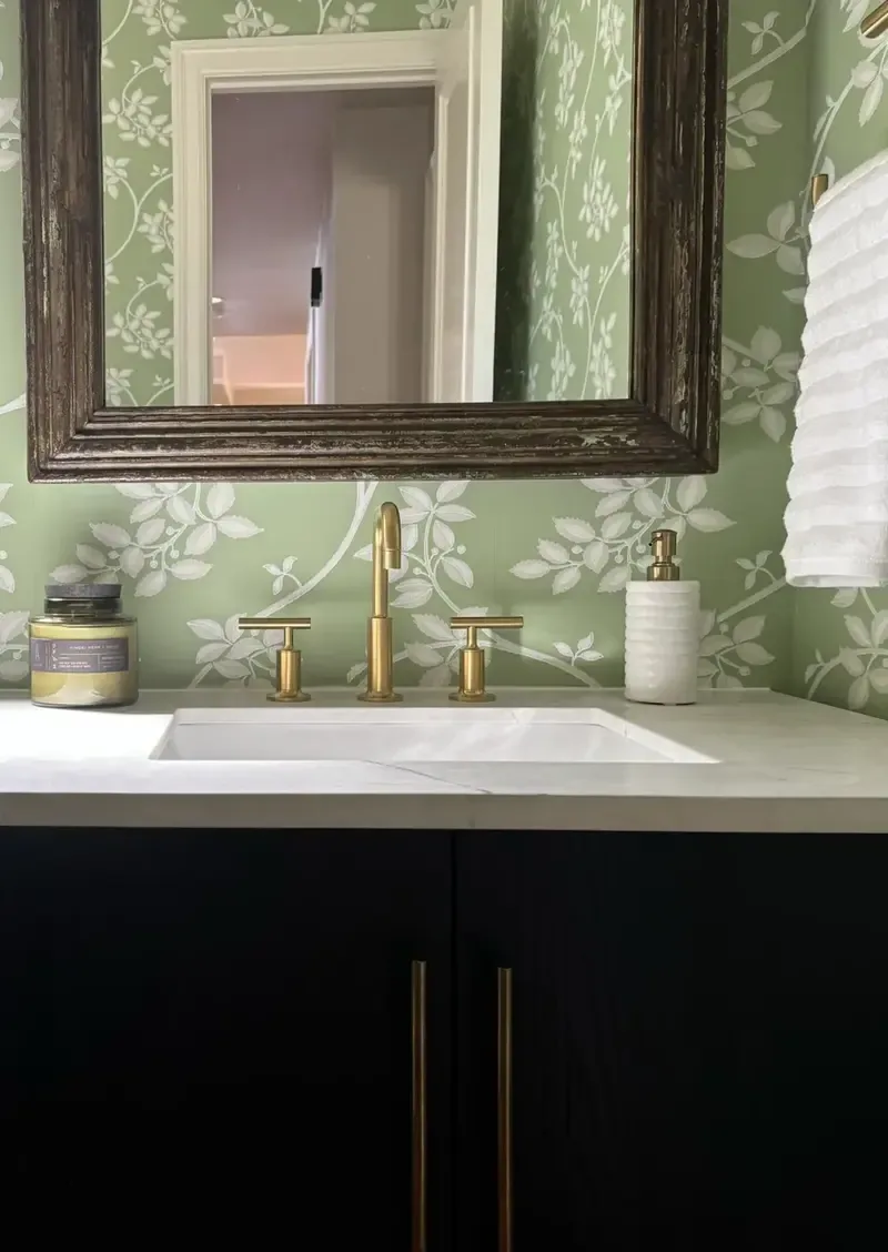 A bathroom vanity with a white countertop, gold faucet, dark cabinetry, floral green wallpaper, and a large framed mirror