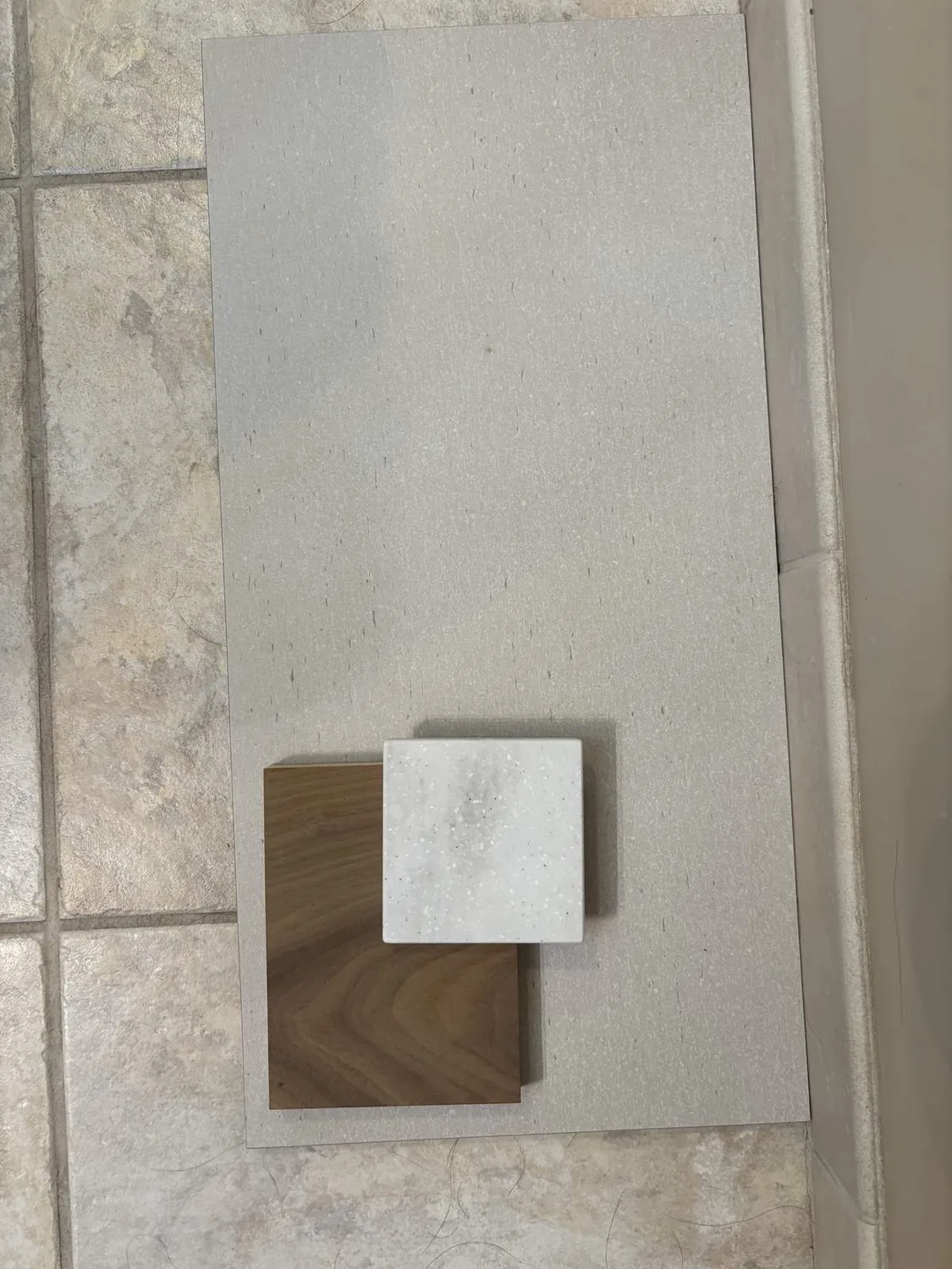 Three material samples including a large light beige tile, a medium wood panel, and a small white speckled square sample arranged on a tiled floor