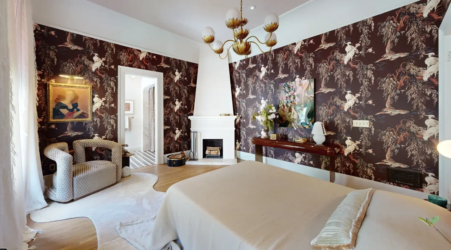 A bedroom with dark floral wallpaper, a white fireplace, a modern armchair, and a bed with neutral bedding