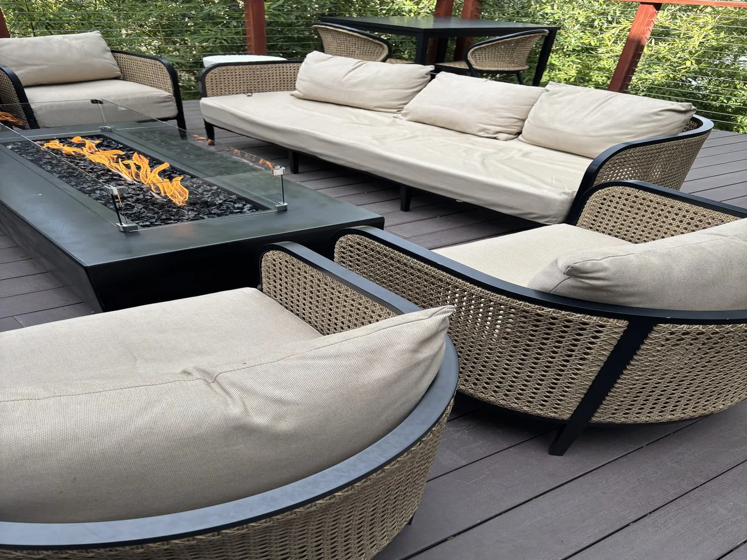 Outdoor seating area with beige cushioned wicker furniture surrounding a modern fire pit table on a wooden deck