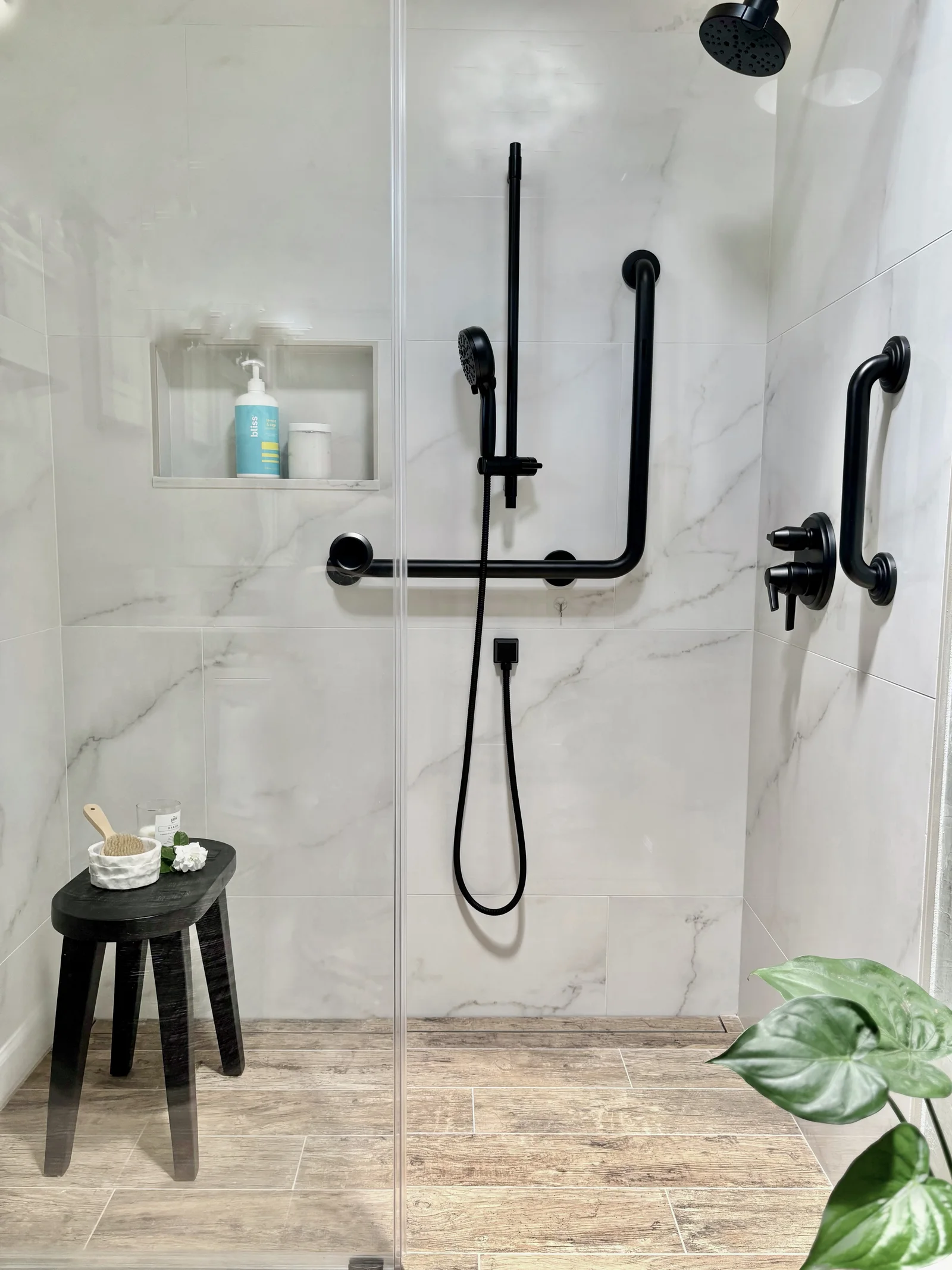 Curbless walk-in shower in Folsom with marble-look large-format porcelain, matte black grab bars and slide-bar hand shower, recessed niche, wood-look plank tile floor continuing into the shower